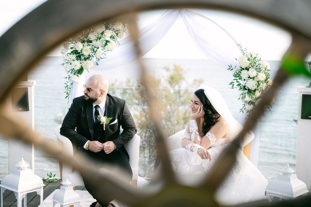 Bride and groom exchanging vows under a floral arch at an outdoor wedding ceremony, framed by a decorative wheel.