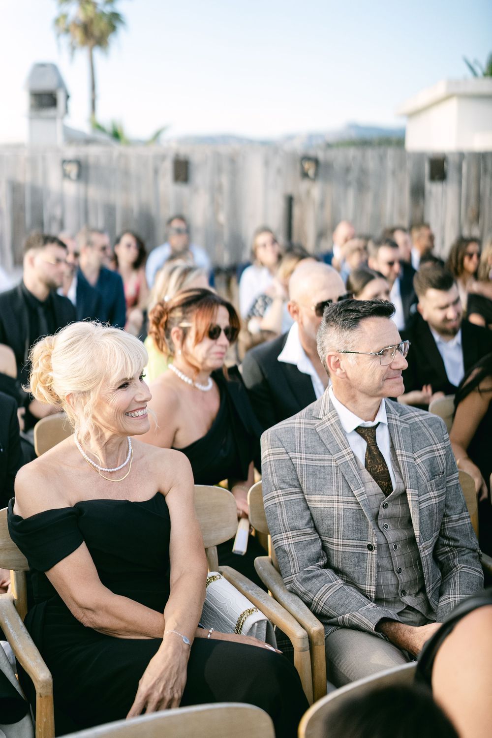 Elegantly dressed wedding guests seated outdoors, smiling woman in black dress with pearls beside man in plaid suit.