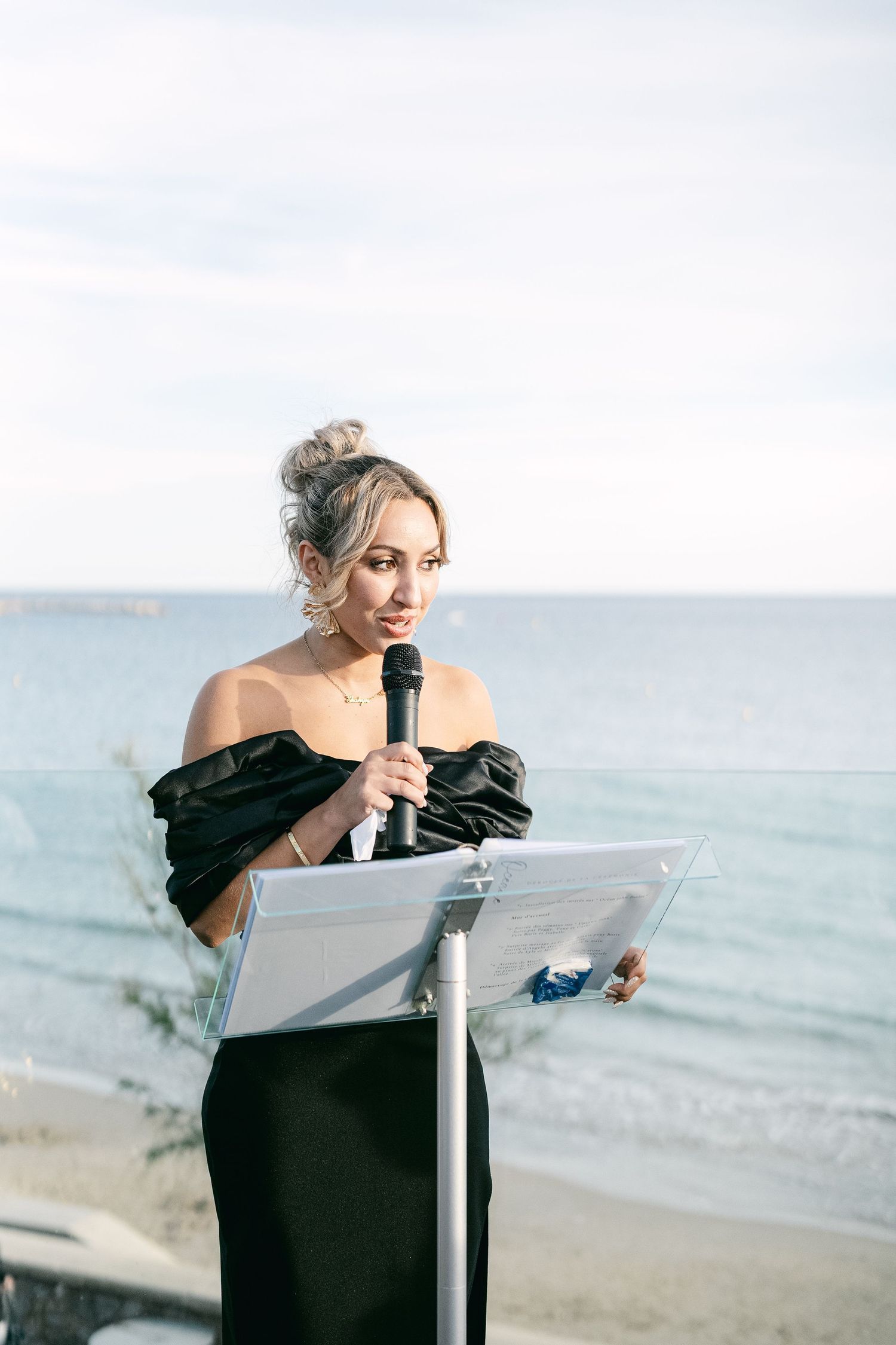 Woman in black off-shoulder dress speaking at podium with microphone by the ocean.