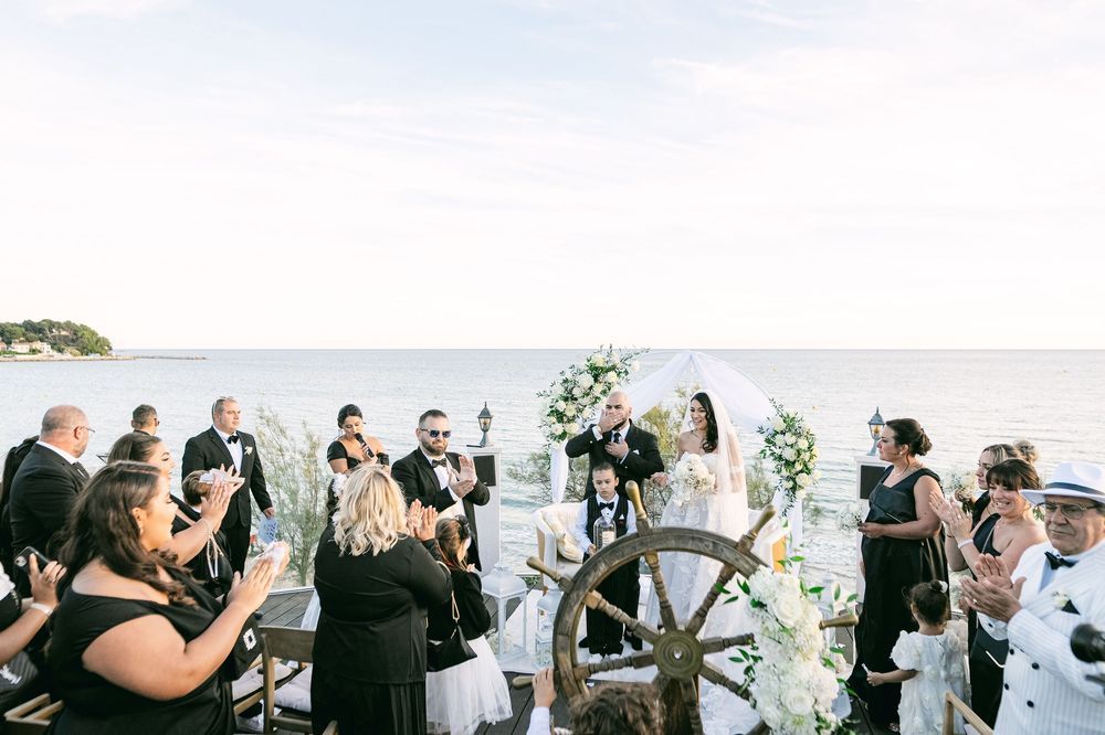 Outdoor waterfront wedding ceremony with couple at floral arch, guests applauding, and a decorative ship wheel in foreground.