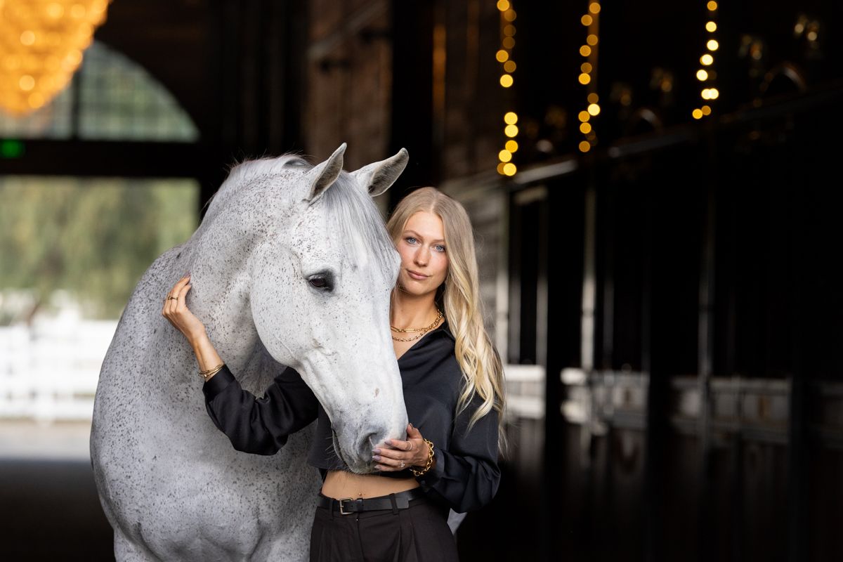 Blonde woman in a black outfit stands affectionately beside a dapple gray horse inside a dimly lit stable with warm string lights.