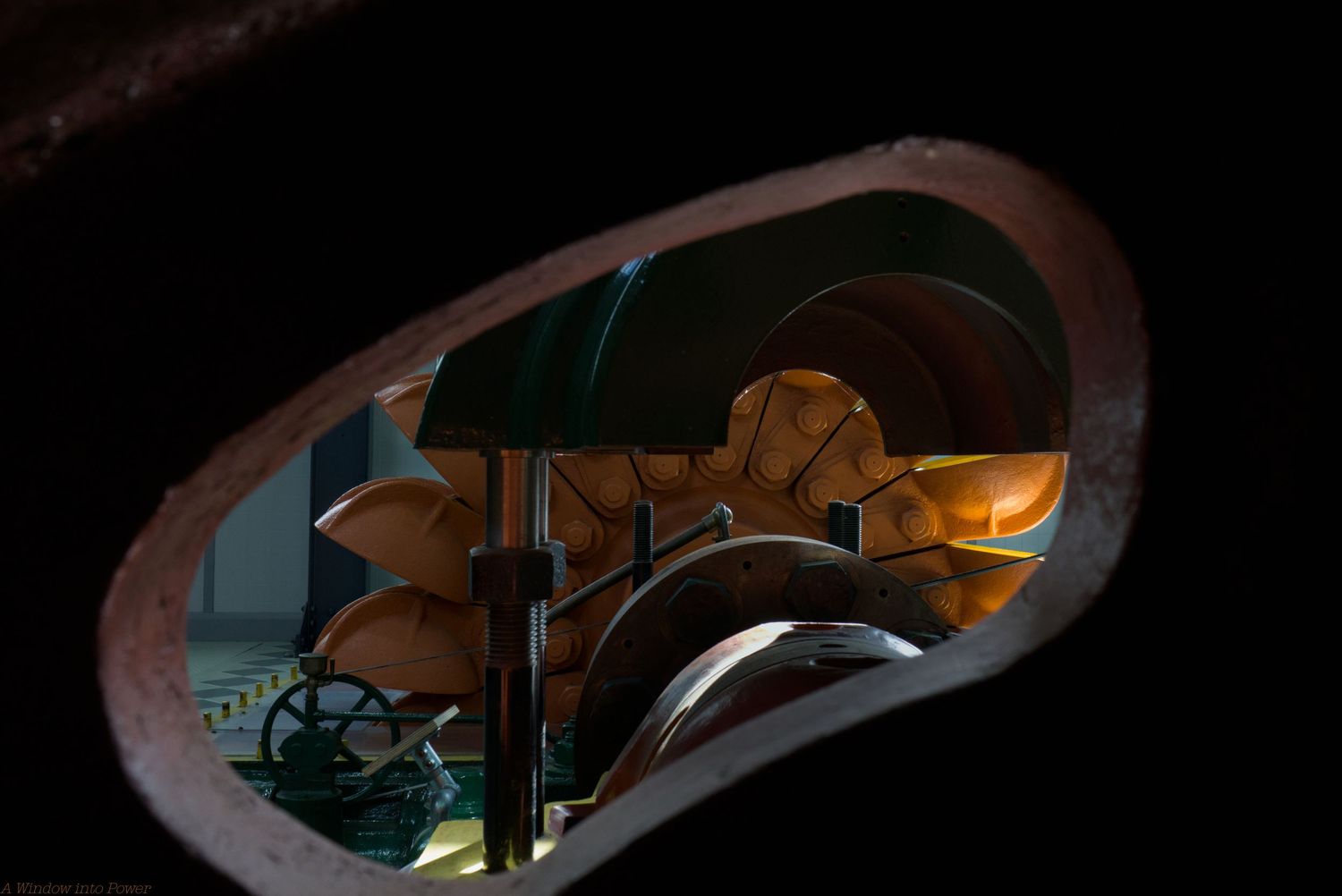 View through an oval opening of a vintage Pelton water turbine wheel with curved copper buckets and mechanical gears in dim light.