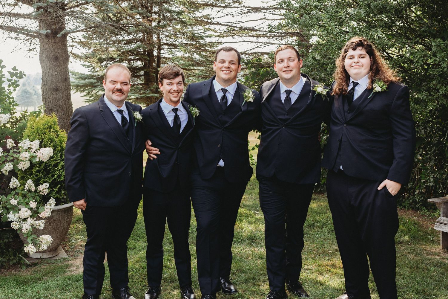 Groom and four groomsmen posing together in black suits beneath evergreen trees.