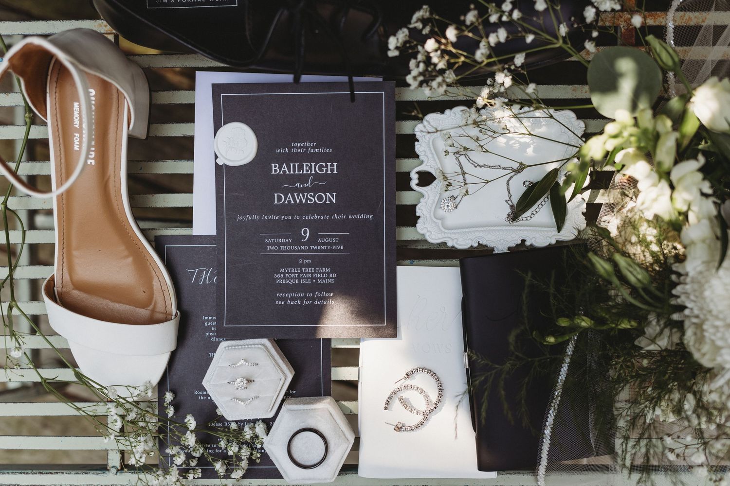 Wedding invitation, white bridal shoe, rings, jewelry, and greenery arranged together on a slatted surface.