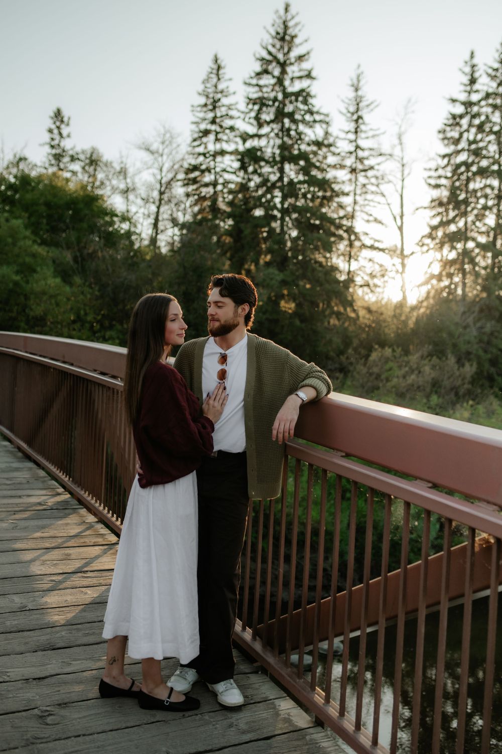 Couple embracing on a wooden bridge at golden hour, surrounded by evergreen trees, in a romantic engagement photo session.