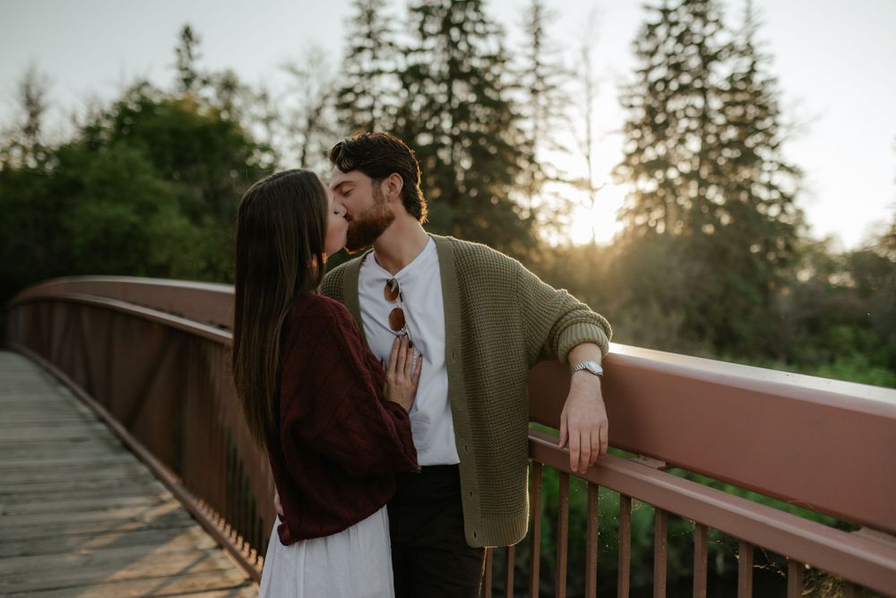 Couple in cozy sweaters share an intimate moment on a wooden bridge at golden hour, surrounded by tall evergreen trees.