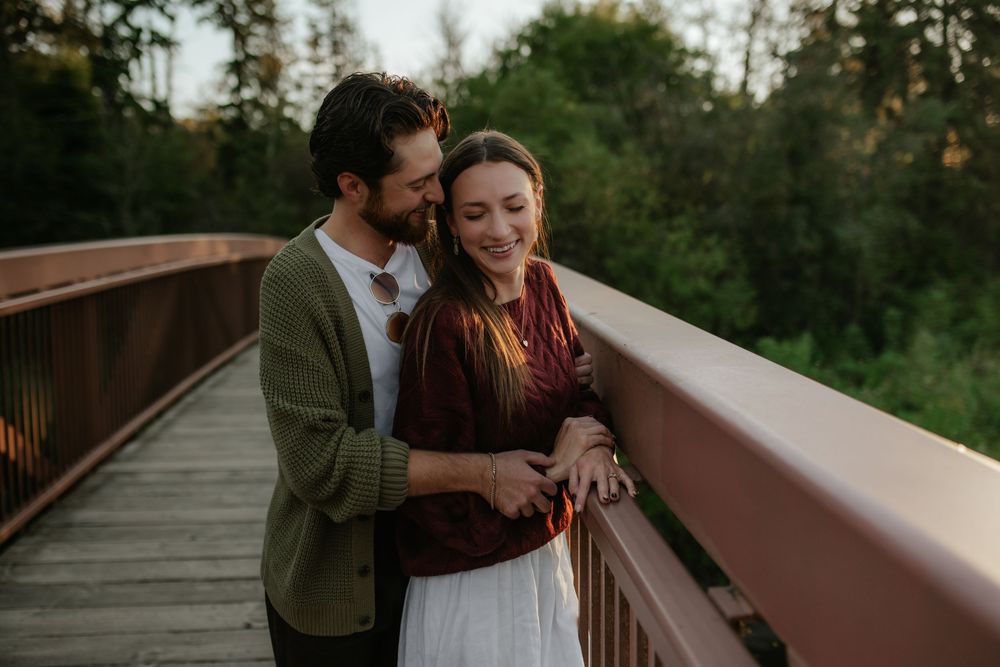 Smiling couple embracing on a wooden footbridge at sunset, the man hugging the woman from behind with trees in the background.