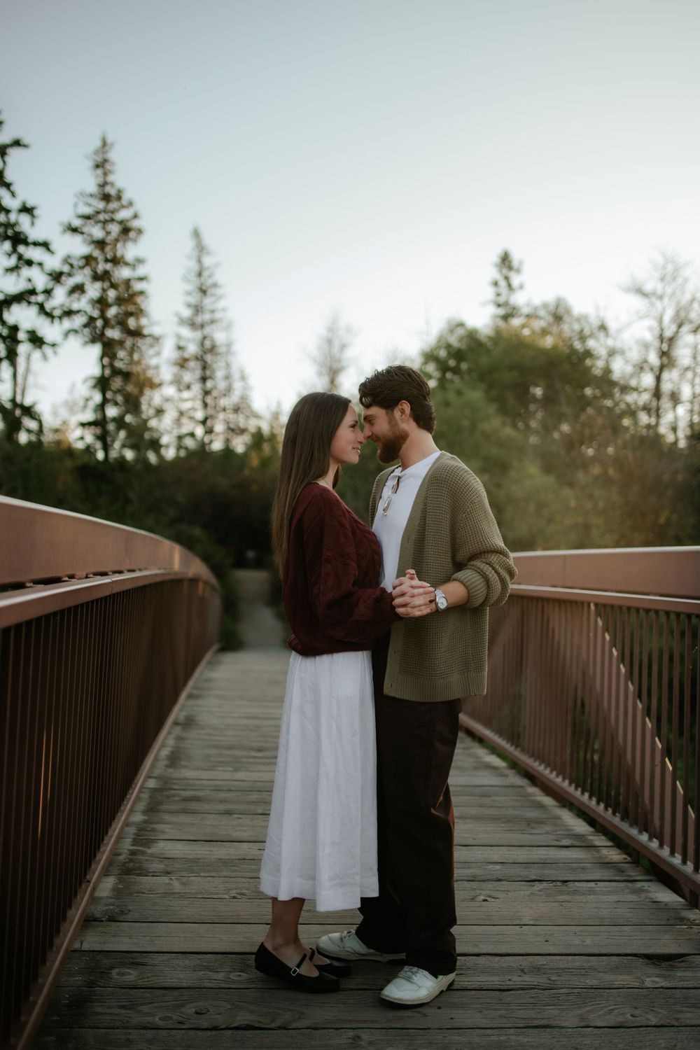 Romantic couple embracing forehead to forehead on a wooden bridge surrounded by evergreen trees during an outdoor engagement photo session.