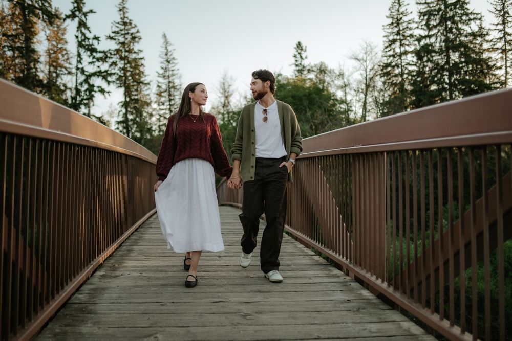 Couple holding hands and walking across a wooden footbridge surrounded by evergreen trees at sunset.