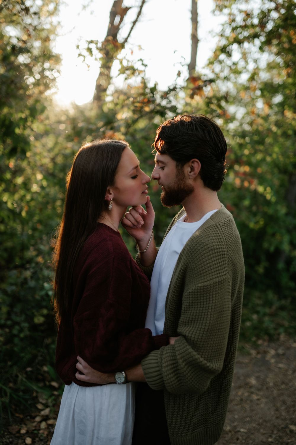 Couple embracing in a sunlit forest as she touches his face, both in cozy autumn sweaters, during an engagement photo session.