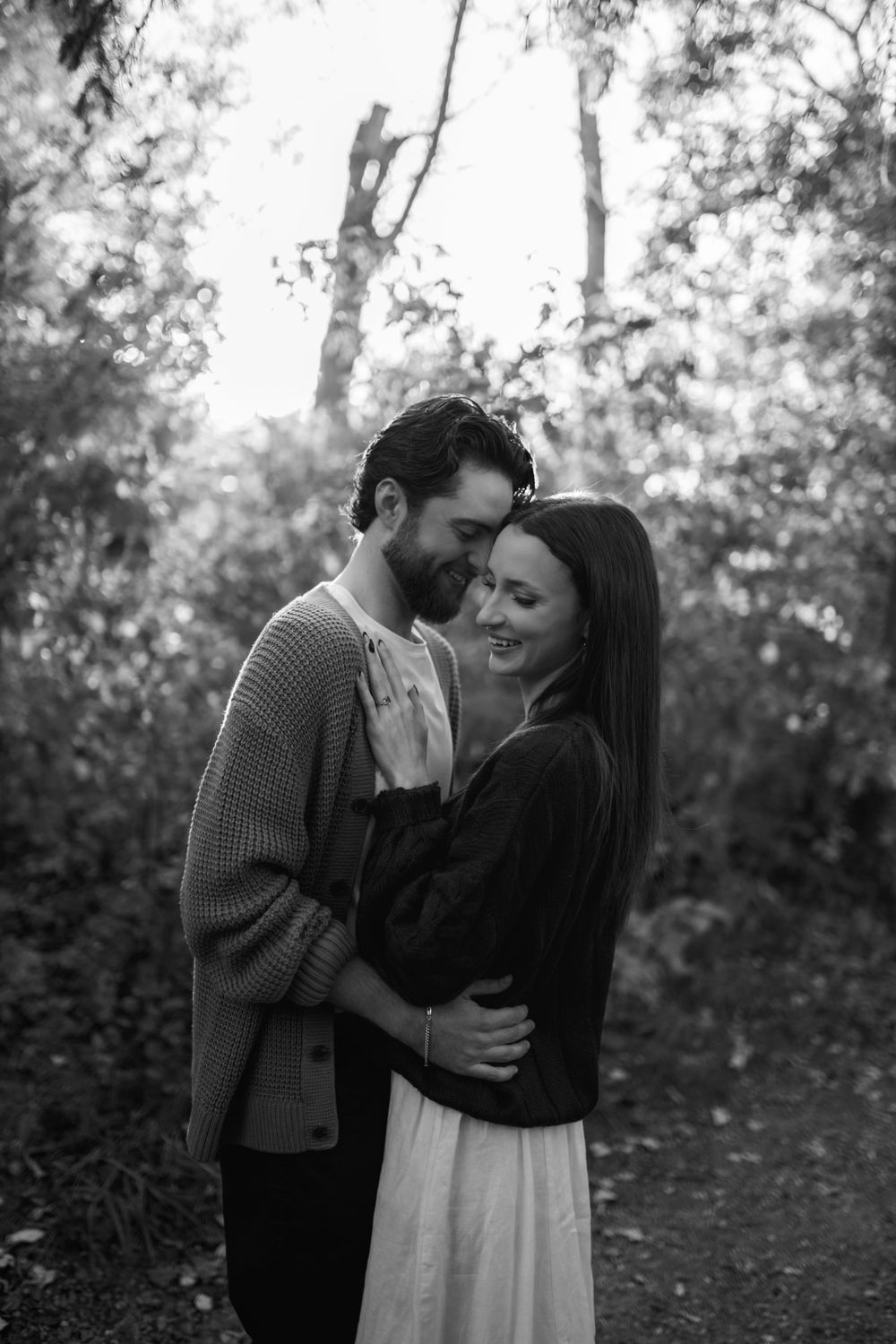 Black and white photo of a smiling couple embracing in a wooded setting, foreheads touching during an outdoor engagement session.