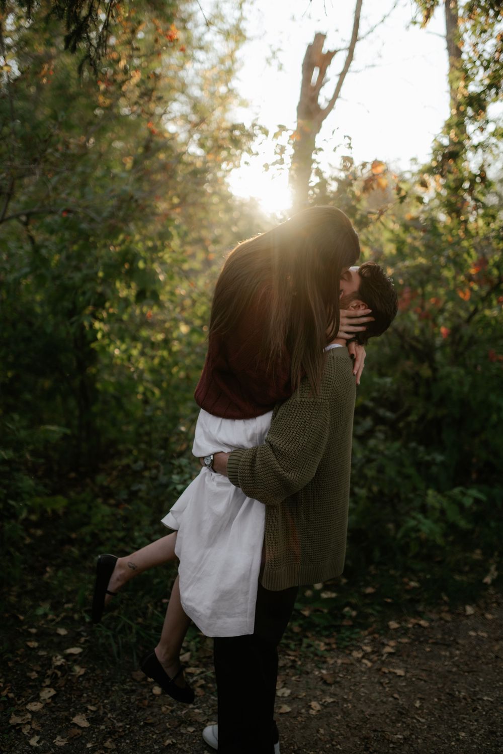 A man lifts a woman in a white dress into an embrace as golden sunlight filters through the trees of a forest path.