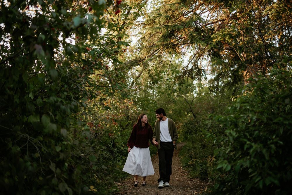 A couple holds hands while walking along a leafy forest path surrounded by lush green foliage during an outdoor engagement photo session.