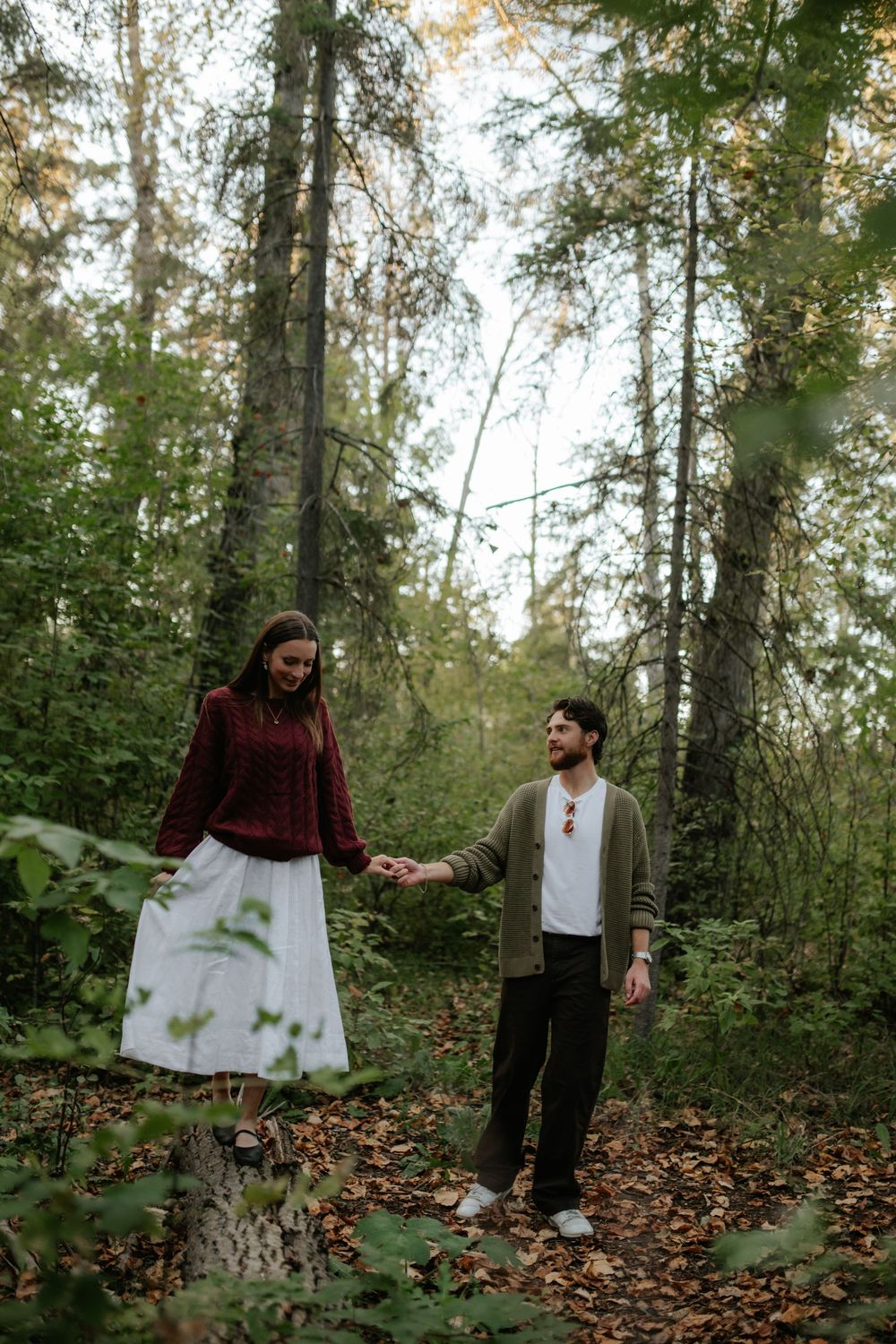 A couple holds hands while walking along a leaf-covered path through a sunlit forest, she in a burgundy sweater and white skirt.