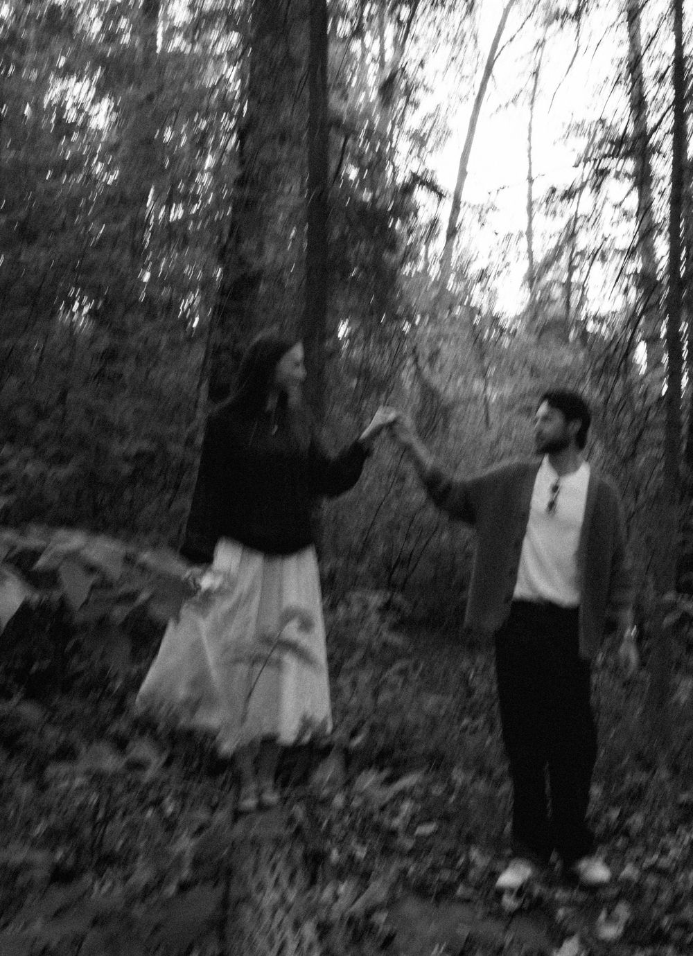 Black and white photo of a couple holding hands in a misty forest, she in a long skirt and he in a cardigan, surrounded by tall trees.