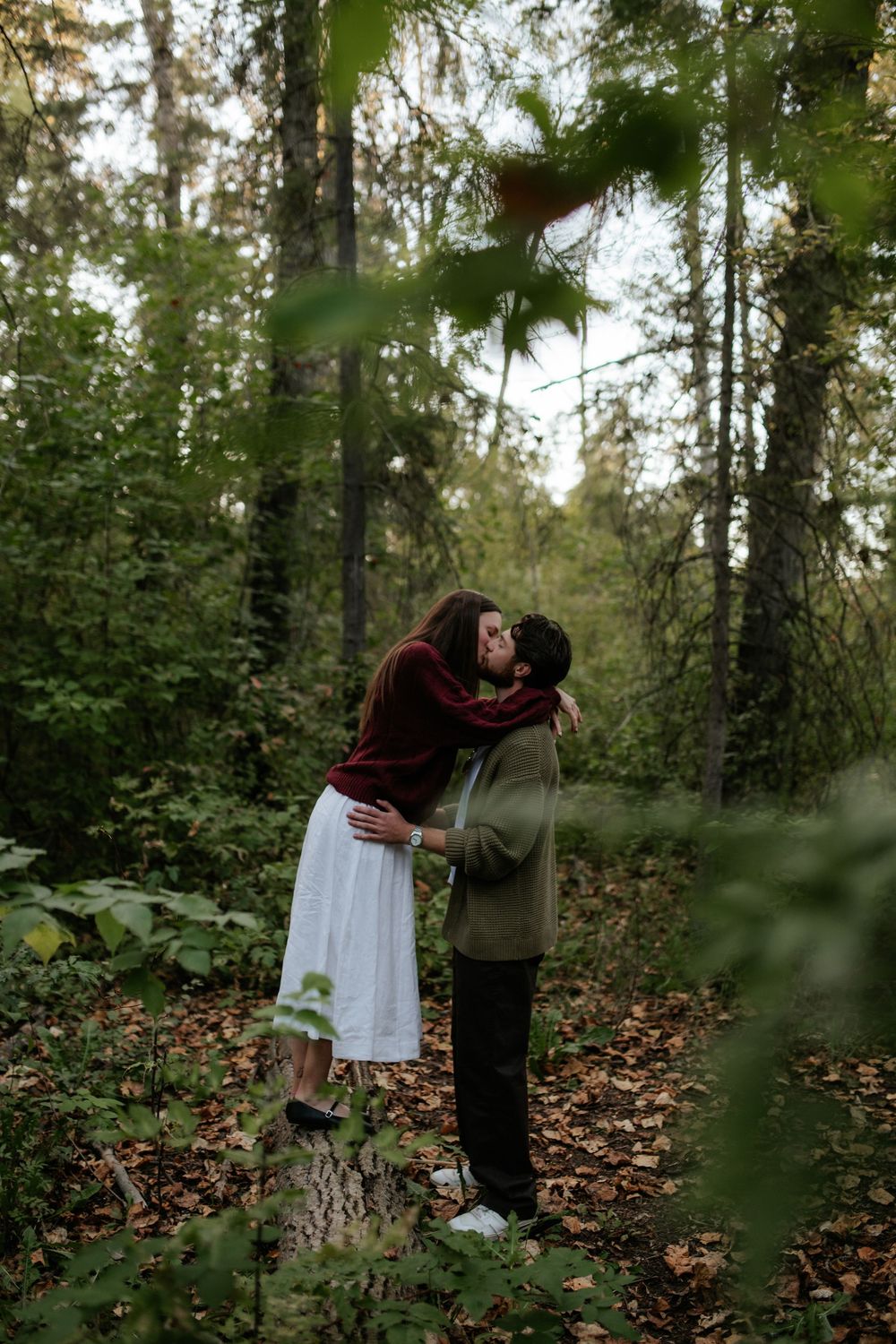 Woman in a burgundy sweater and white skirt stands on a tree stump kissing a man in an olive cardigan in an autumn forest.