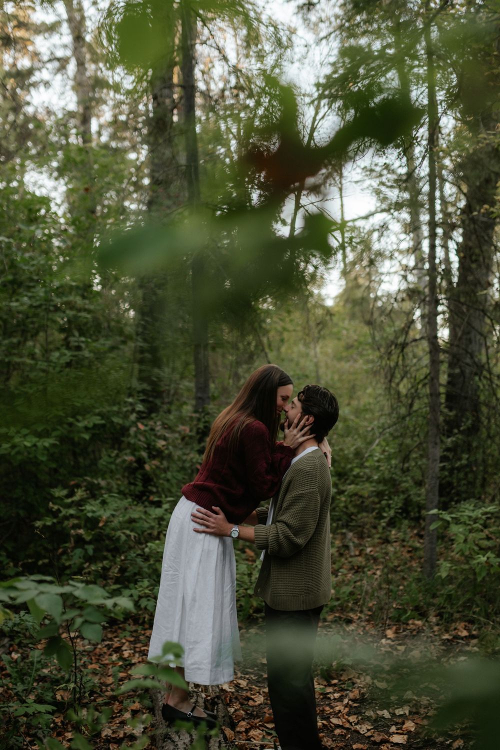 A couple shares a kiss on a leaf-covered woodland trail, surrounded by tall trees and green foliage.