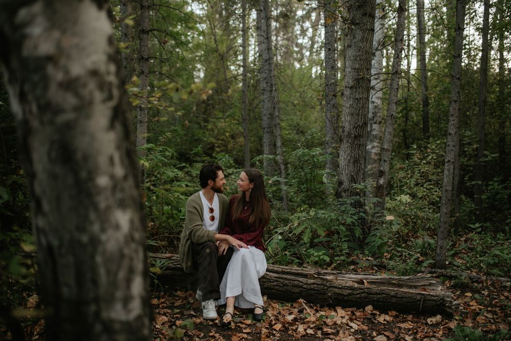Couple sitting close together on a fallen log in a moody, tree-filled forest, sharing an intimate moment surrounded by autumn leaves.