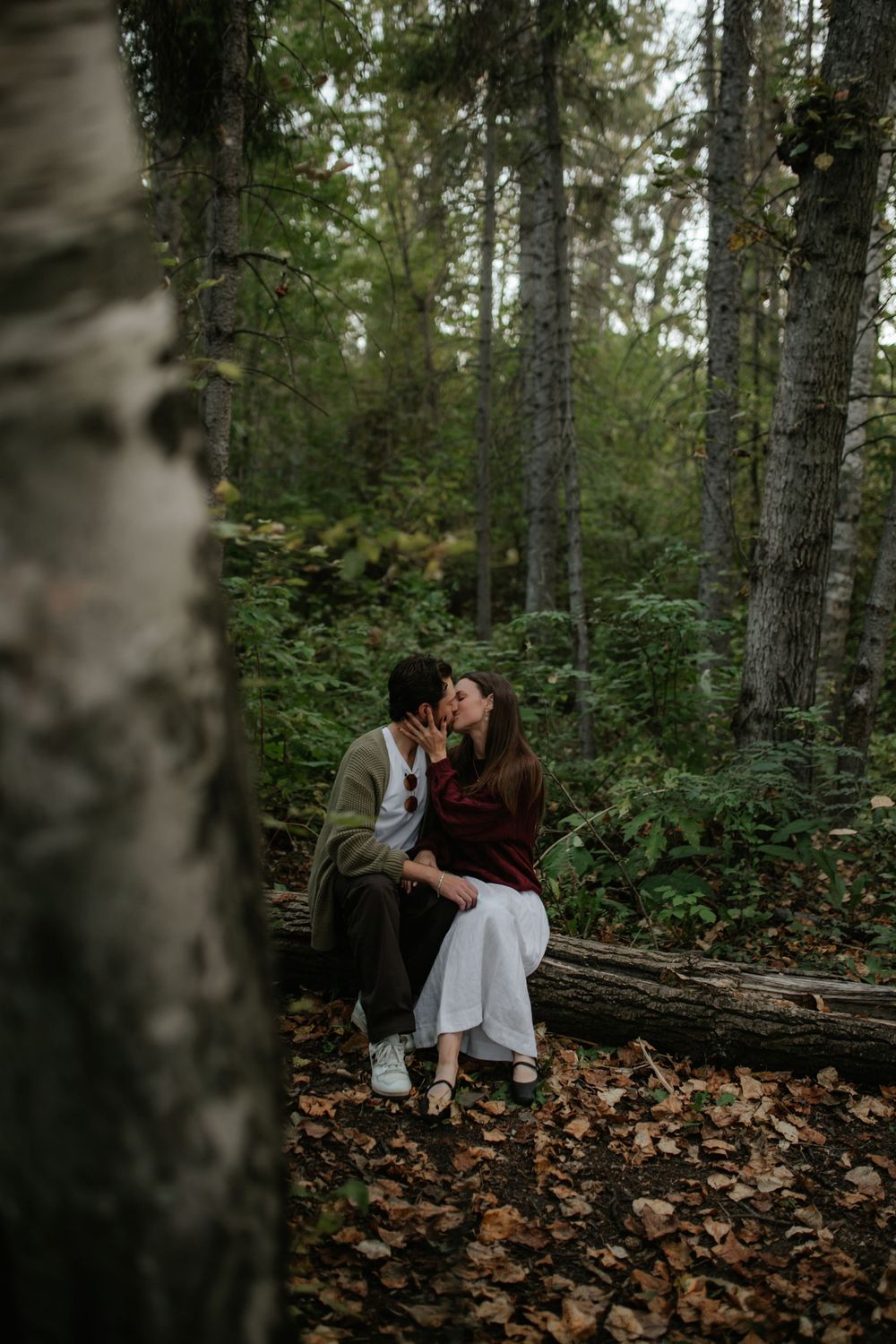 Couple sharing a kiss while seated on a fallen log in an autumn forest surrounded by tall trees and fallen leaves.
