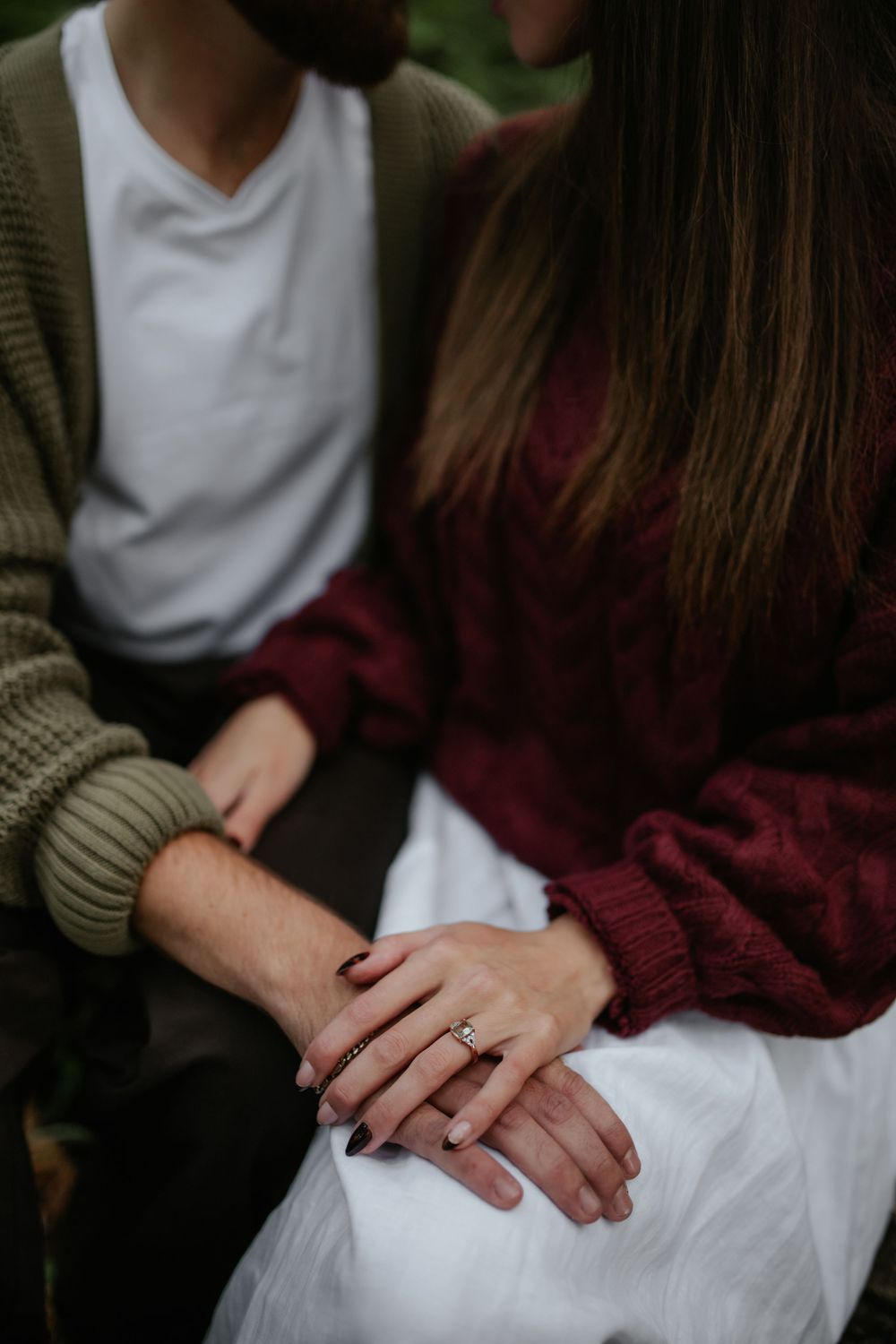 Close-up of a seated couple holding hands, her ring visible, she in a burgundy knit sweater and white skirt, he in a green cardigan.