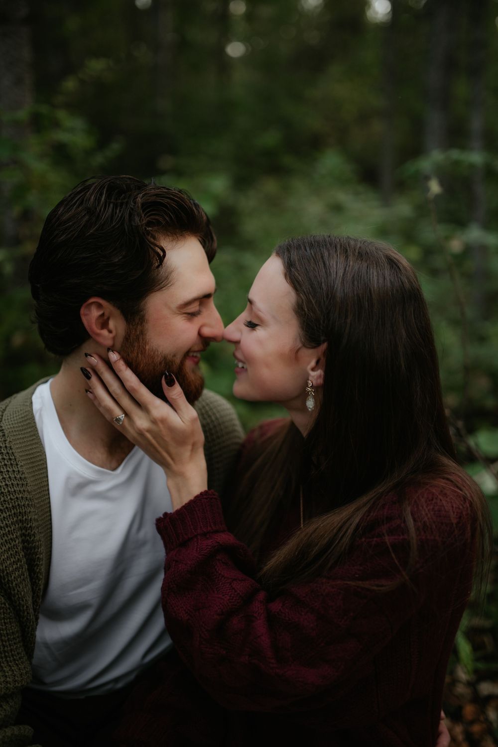 Close-up of a bearded man and a long-haired woman nose to nose, smiling in a romantic moment among green forest trees.