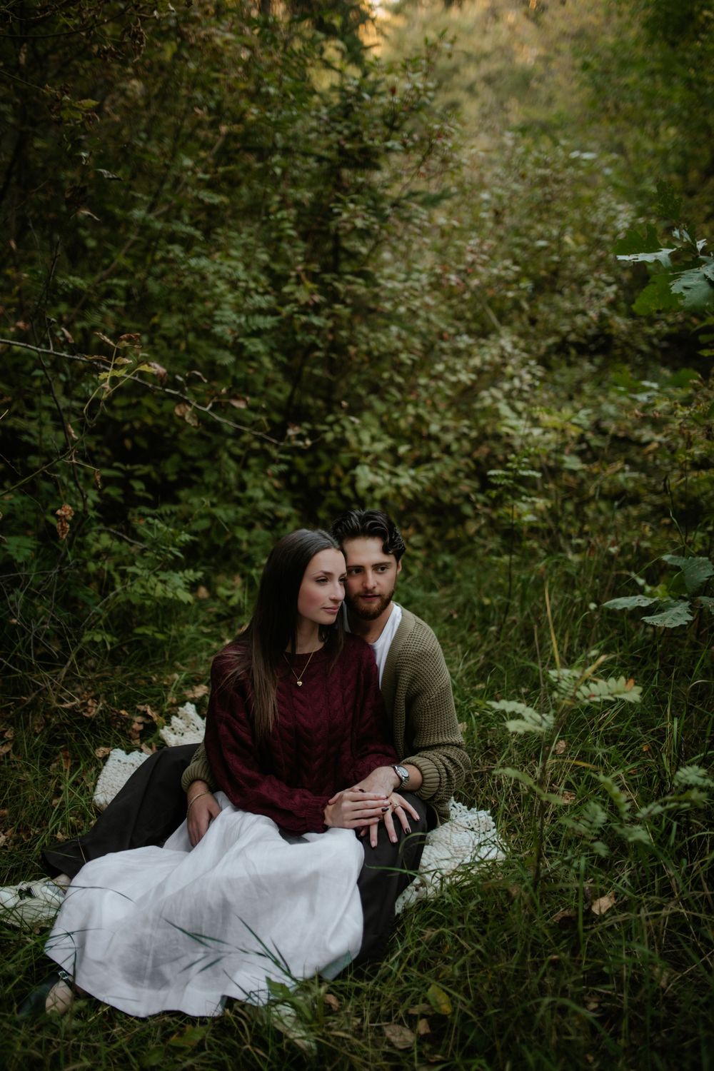 Couple sitting close together on a blanket in a lush green forest, she wears a burgundy sweater and white skirt for an engagement photo.