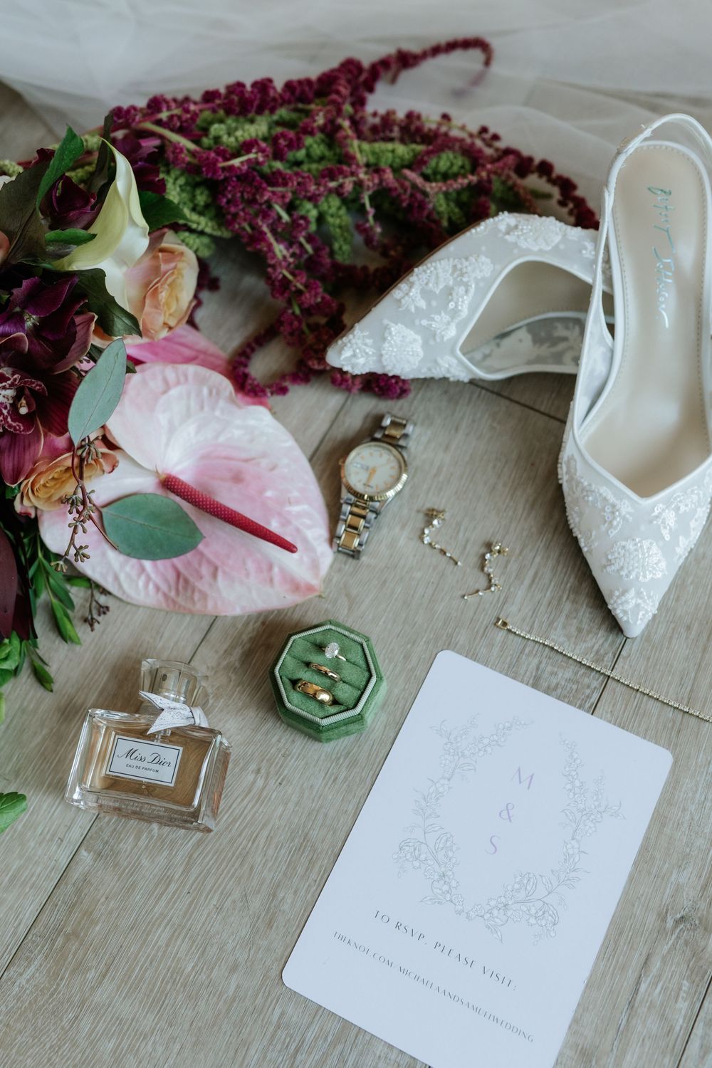 Bridal flat lay with lace wedding heels, burgundy and pink floral bouquet, gold watch, perfume bottle, green ring box and invitation card.