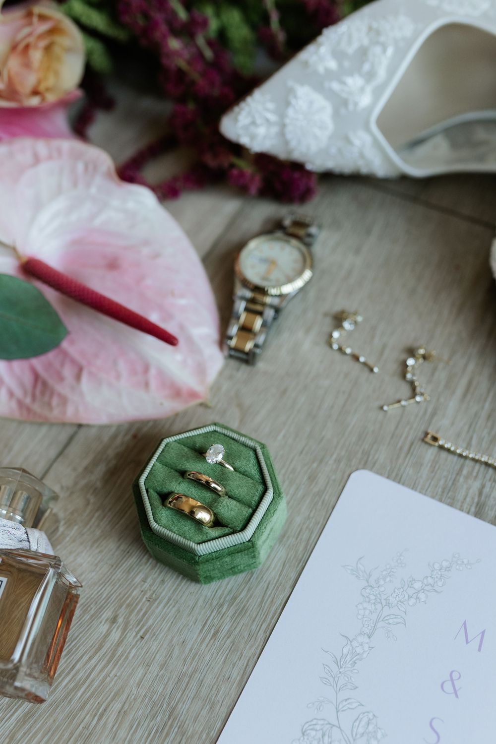 Green velvet hexagonal ring box holding wedding bands and a diamond ring beside a watch, earrings, perfume, bridal shoe, and pink anthurium.