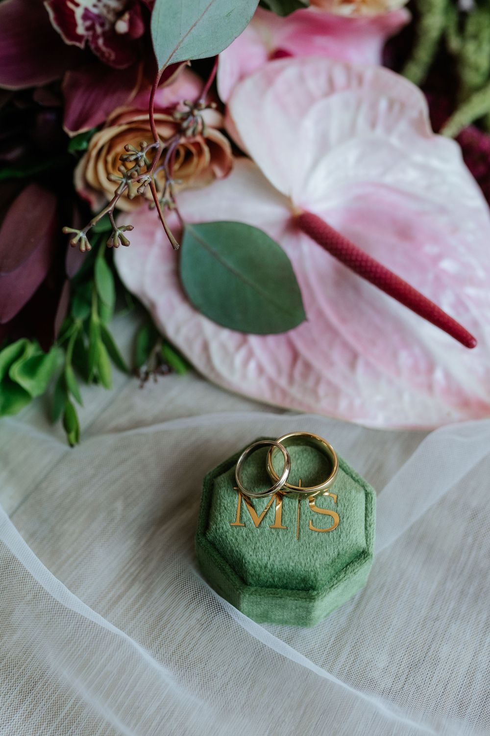Two gold wedding rings rest in a green velvet ring box monogrammed MJ beside pink anthurium and burgundy bridal bouquet flowers.