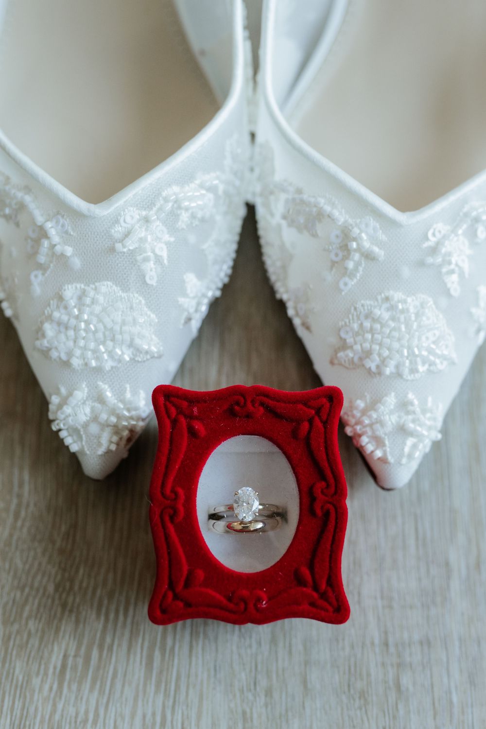Pointed-toe white bridal flats with beaded floral lace appliqués beside a solitaire diamond engagement ring in a red velvet box.