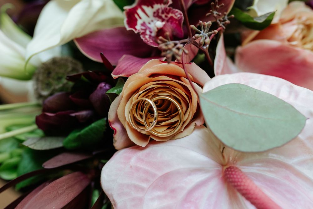 Close-up of a wedding bouquet with a gold ring resting in a tan rose, surrounded by pink anthurium, burgundy blooms and eucalyptus leaves.