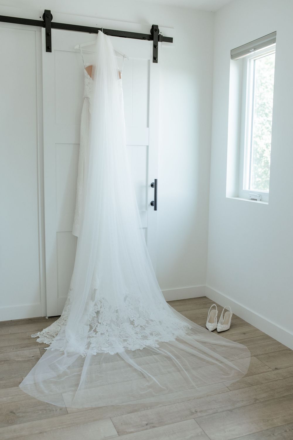White lace wedding gown and long tulle veil hanging on a barn door beside white bridal heels in a bright room.