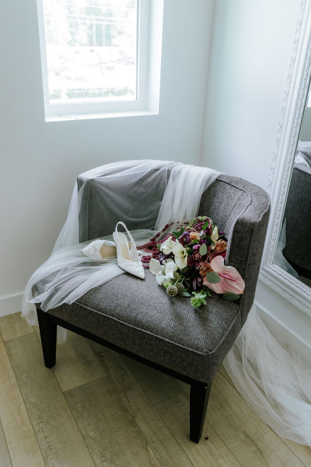 Bridal bouquet, white heels, and a flowing veil rest on a gray accent chair beside a window and ornate mirror.