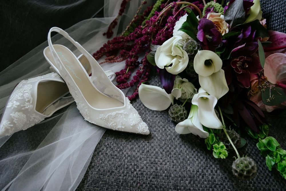 White lace slingback bridal heels beside a sheer veil and a bouquet of white calla lilies, purple blooms and burgundy amaranthus.