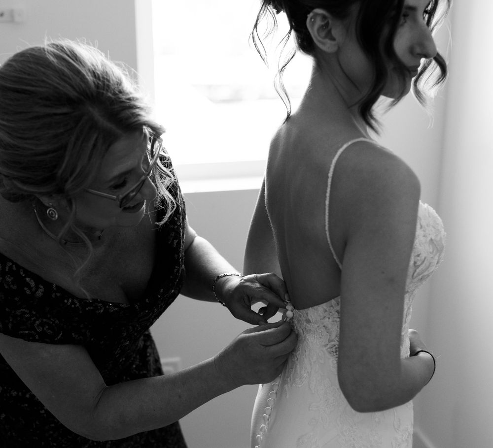 A woman in a dark patterned dress fastens the buttons on the back of a bride's lace wedding gown beside a sunlit window.