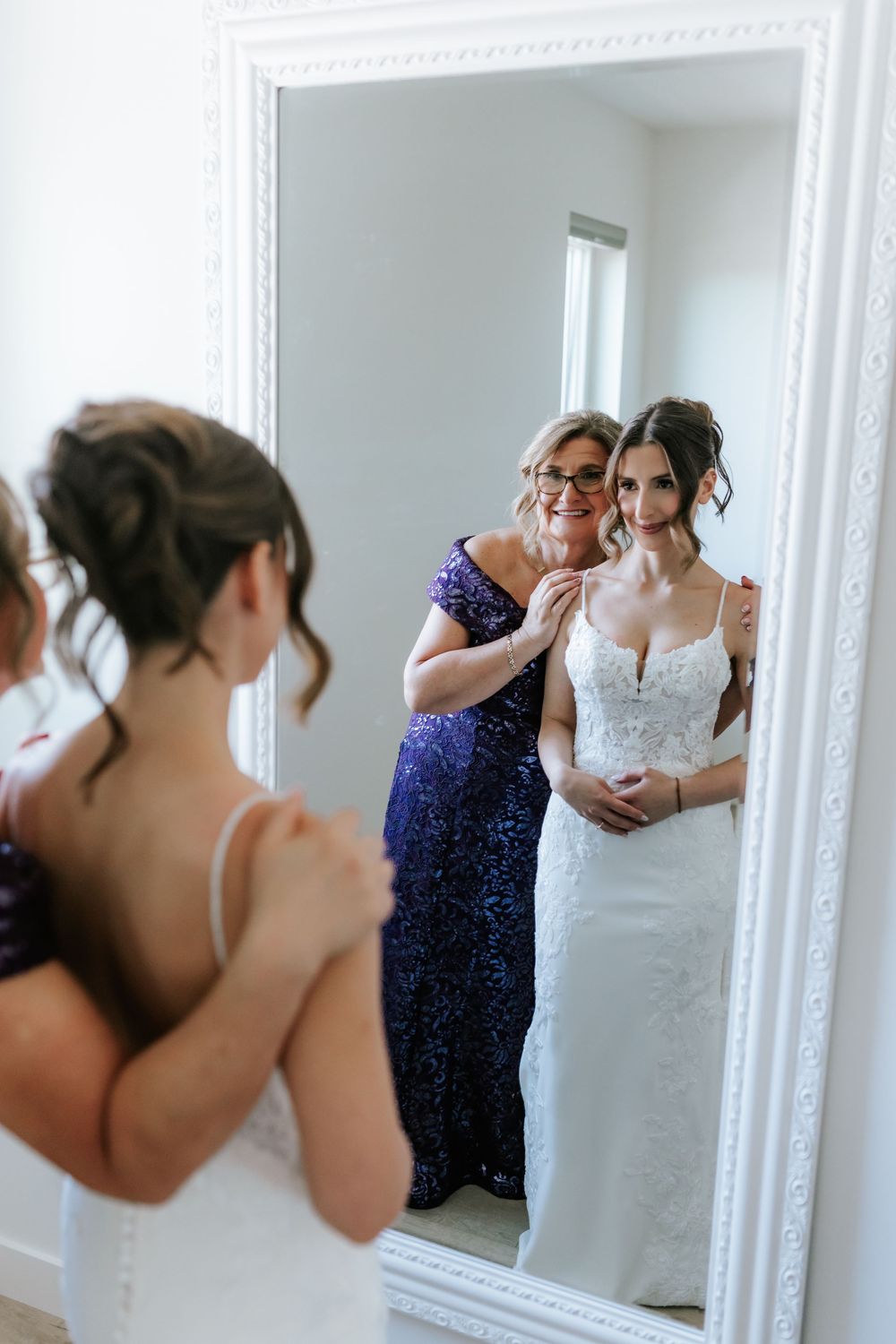A bride in a lace wedding gown smiles in a large white mirror as her mother in a purple dress helps her get ready.