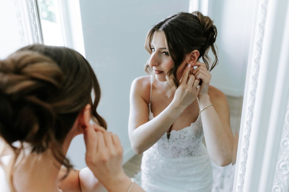 Bride in a white lace wedding dress with an elegant updo adjusts her earring while looking in a mirror in a bright room.