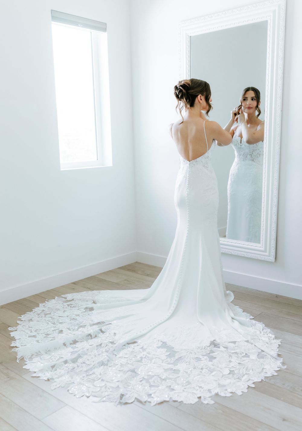 Bride in an elegant lace-trimmed gown with a long train admires herself in a large white-framed mirror in a bright room.