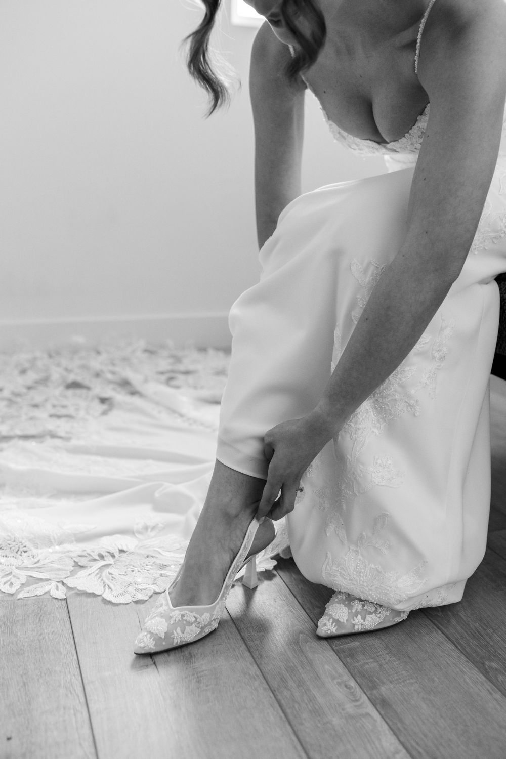 Black and white photo of a bride in a lace wedding gown sitting on a wooden floor while adjusting her lace bridal heel.
