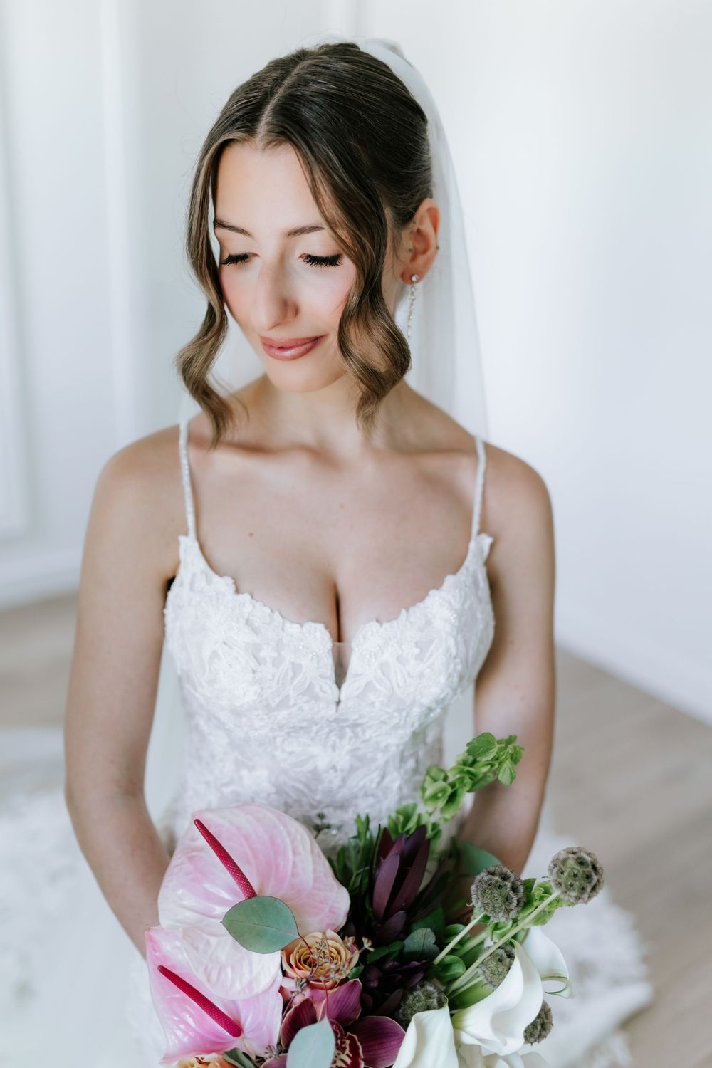 Bride in a lace wedding gown and veil looks down while holding a bouquet of pink anthurium, burgundy blooms and greenery.