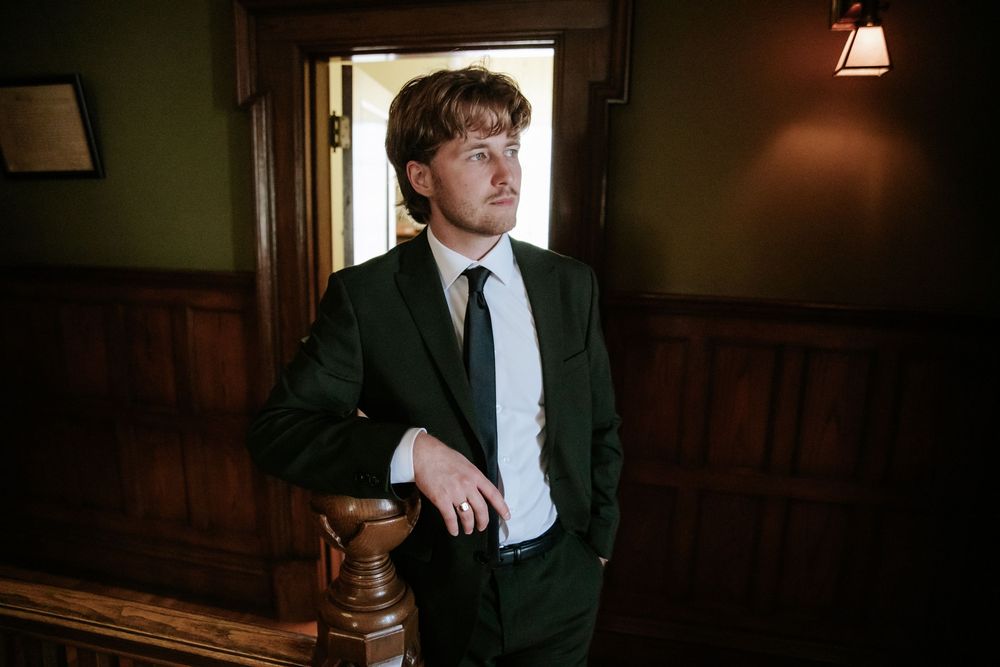 Young man in a dark suit and tie leans against a wooden banister in a warmly lit room with wood paneling.