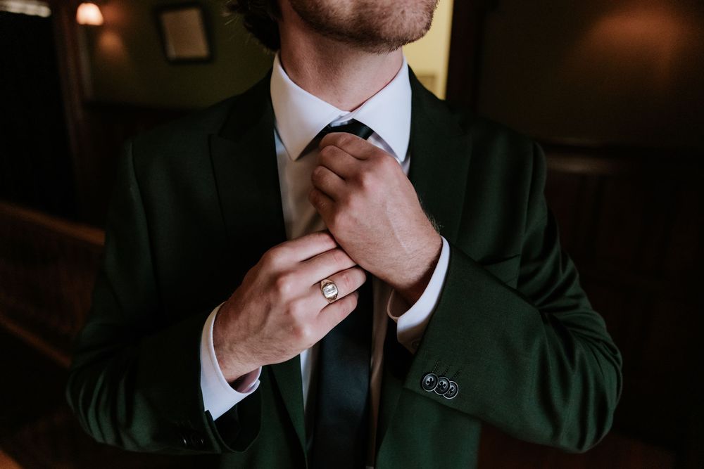 Groom in a dark green suit adjusts his black tie while wearing a ring on his finger.