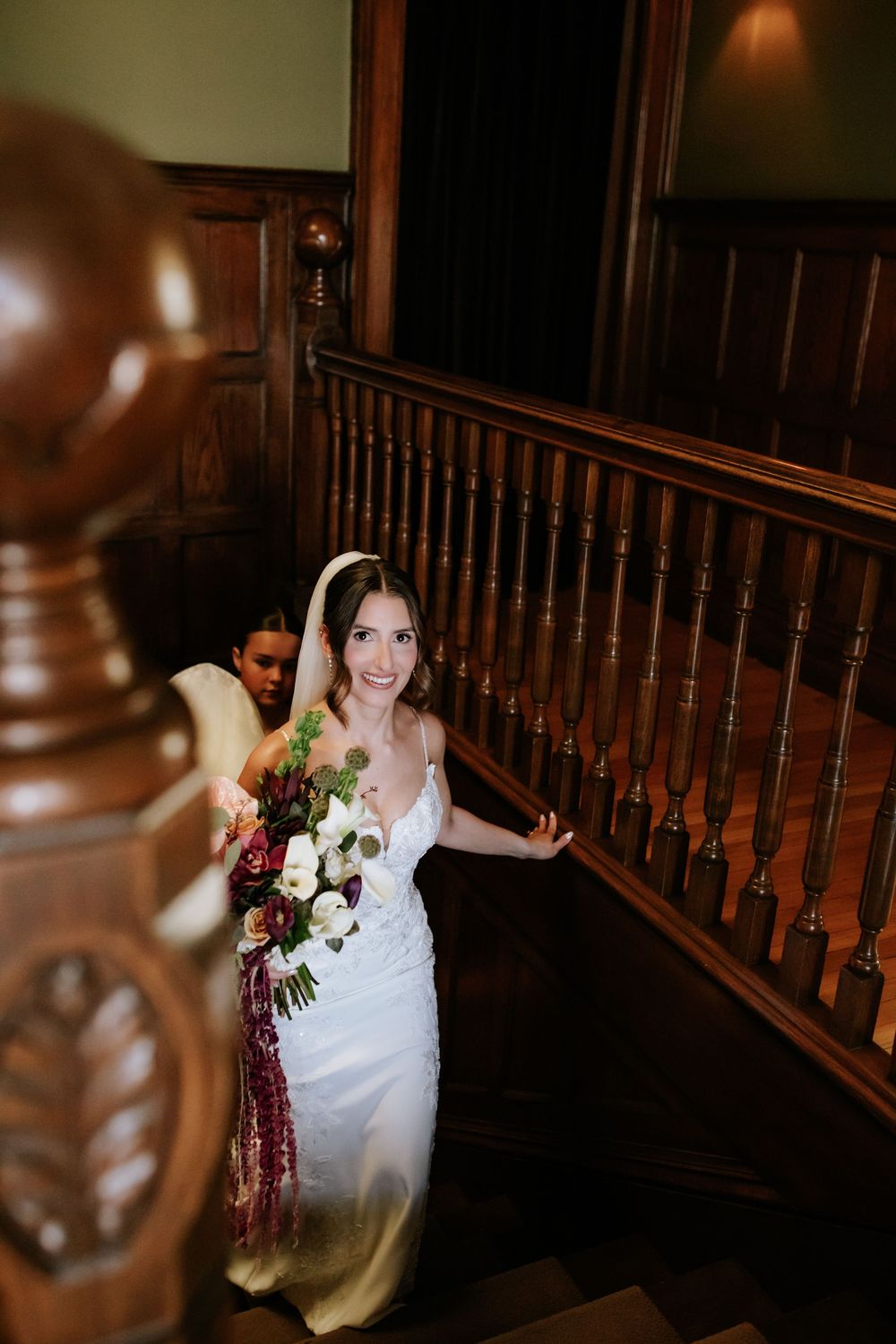Smiling bride in a white satin gown descends a rich wooden staircase holding a lush burgundy and white bouquet with trailing amaranthus.