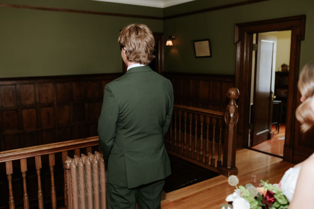 Groom in a green suit stands facing away at the top of a wooden staircase while a bride holding a bouquet waits nearby in a paneled room.