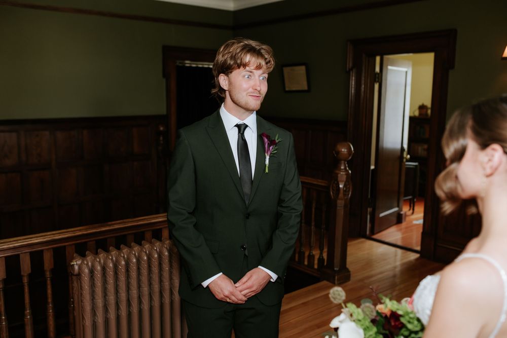 Groom in a dark suit with a boutonniere smiles at a bride holding a bouquet in a wood-paneled room before their wedding ceremony.