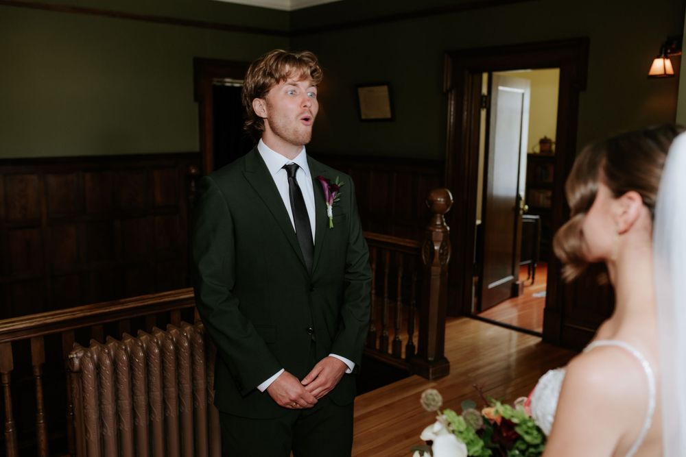 Groom in a dark suit with a boutonniere gasps in awe seeing his bride in a white gown holding a bouquet during a first look.