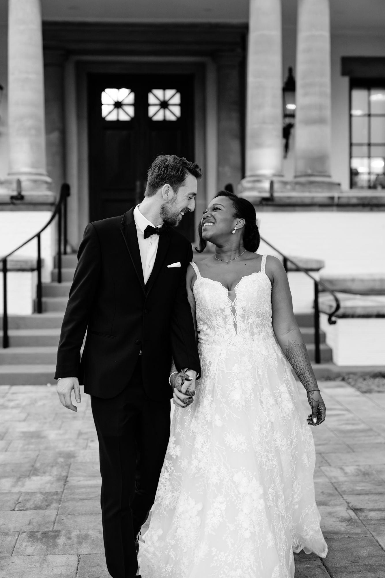 Smiling bride in a lace wedding gown and groom in a tuxedo with bow tie walk hand in hand outside a columned building in black and white.