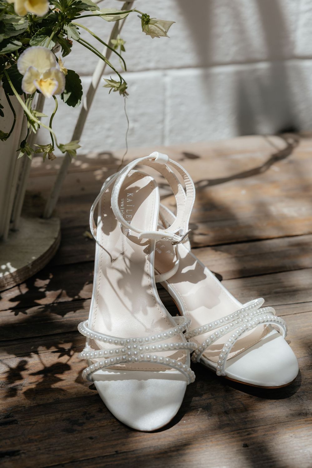White pearl-embellished strappy bridal sandals with ankle straps on a sunlit wooden floor beside white flowers.