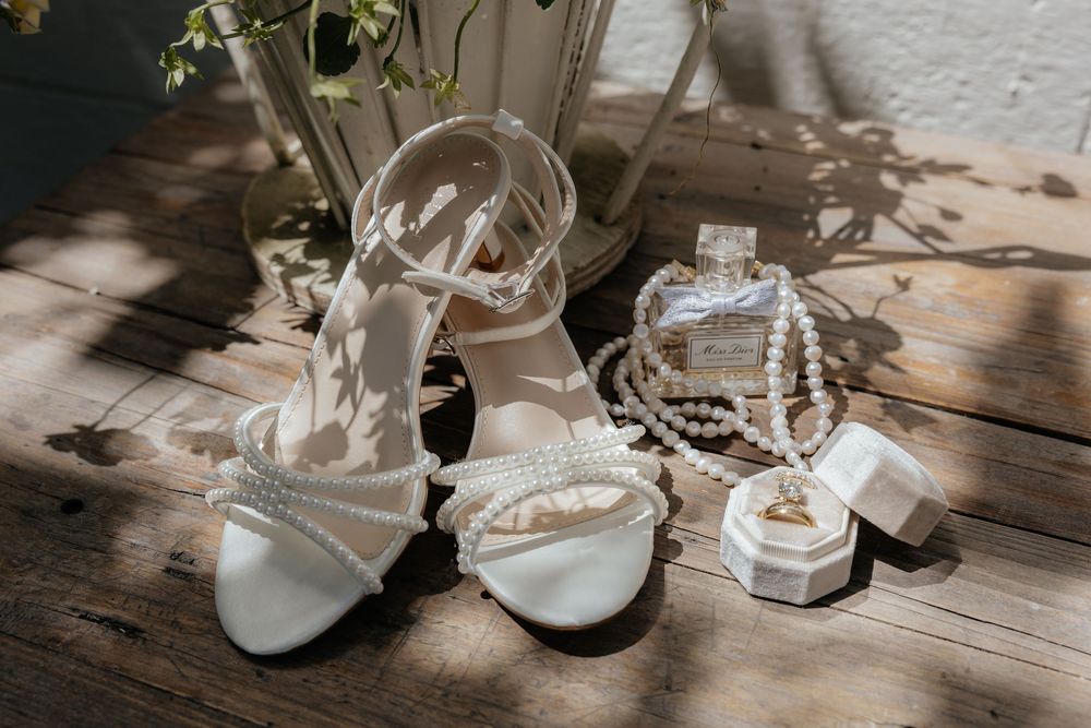 White pearl-embellished strappy bridal sandals beside a pearl necklace, perfume bottle and open ring box on a sunlit rustic wooden surface.