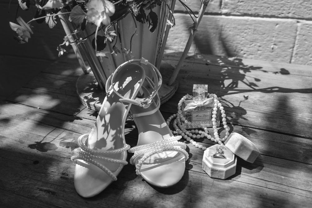 Black and white wedding details of pearl-strap bridal heels, a pearl necklace, perfume bottle and open ring box on a rustic wooden floor.